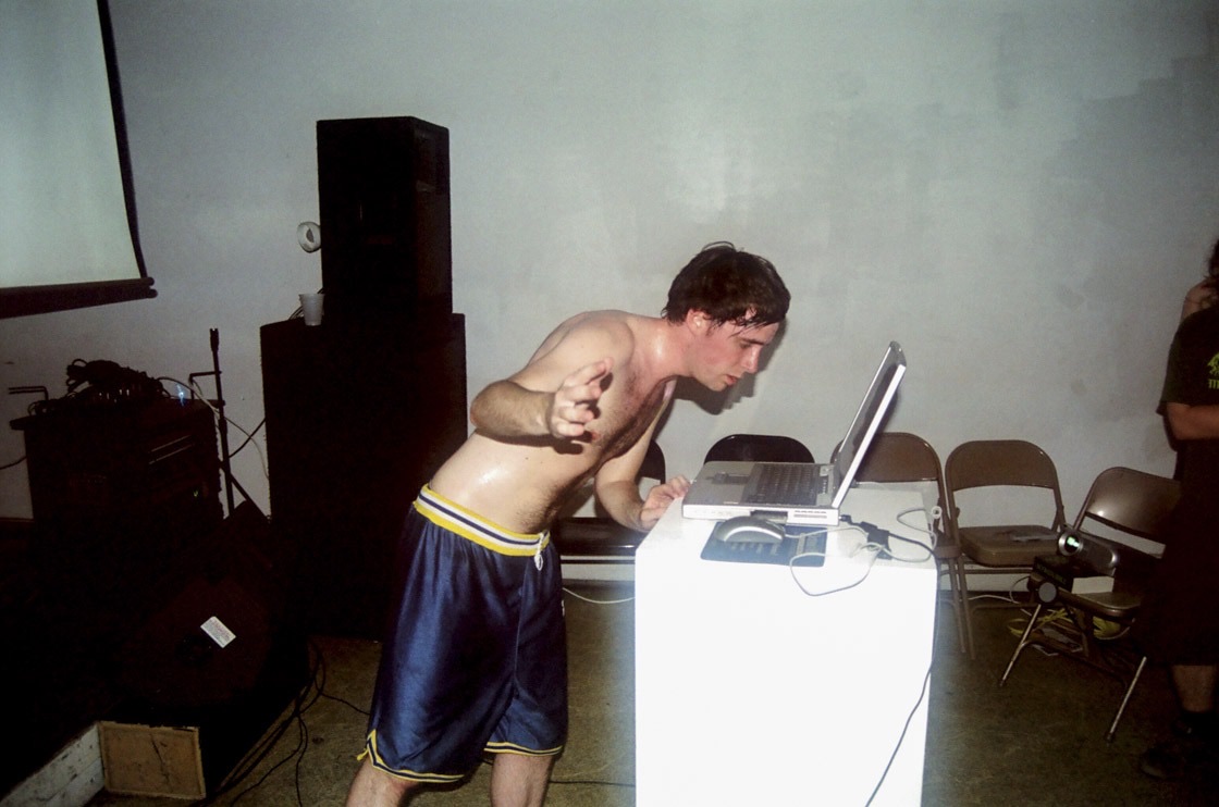 A shirtless, sweaty performer in basketball shorts leans into a laptop on a white plinth, with a speaker stack and a projection screen behind him.